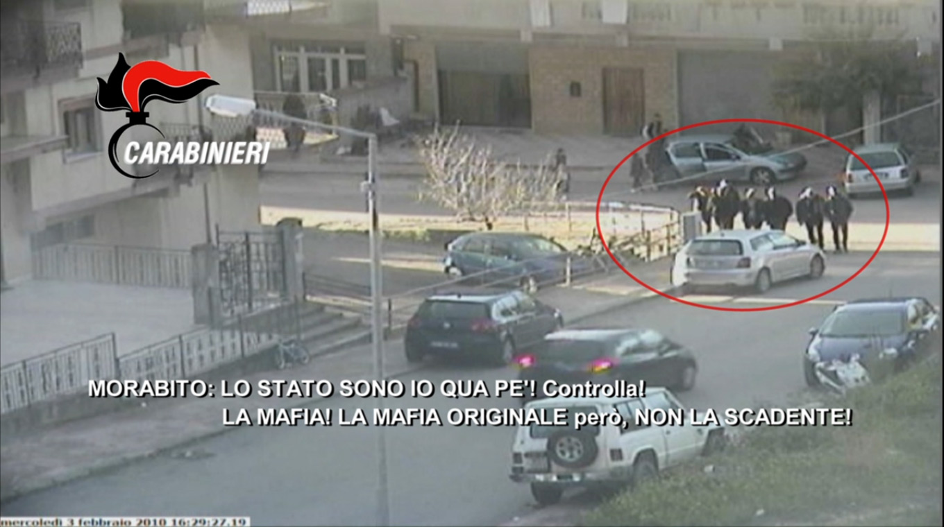 A still shot from a police surveillance video shows 'Ndragheta members walking together. The superimposed wiretrap transcript quotes Rocco Morabito: "The state is me, Pe! Check it! The Mafia! The original Mafia, though, not the shoddy one." (Video credit: Arma dei Carabinieri) 
