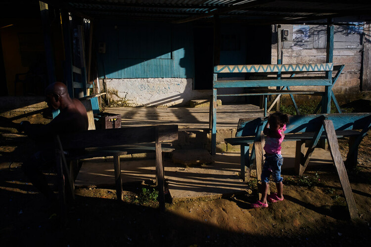 A man sits on a bench, watched by a young girl, in a town in Chocó.