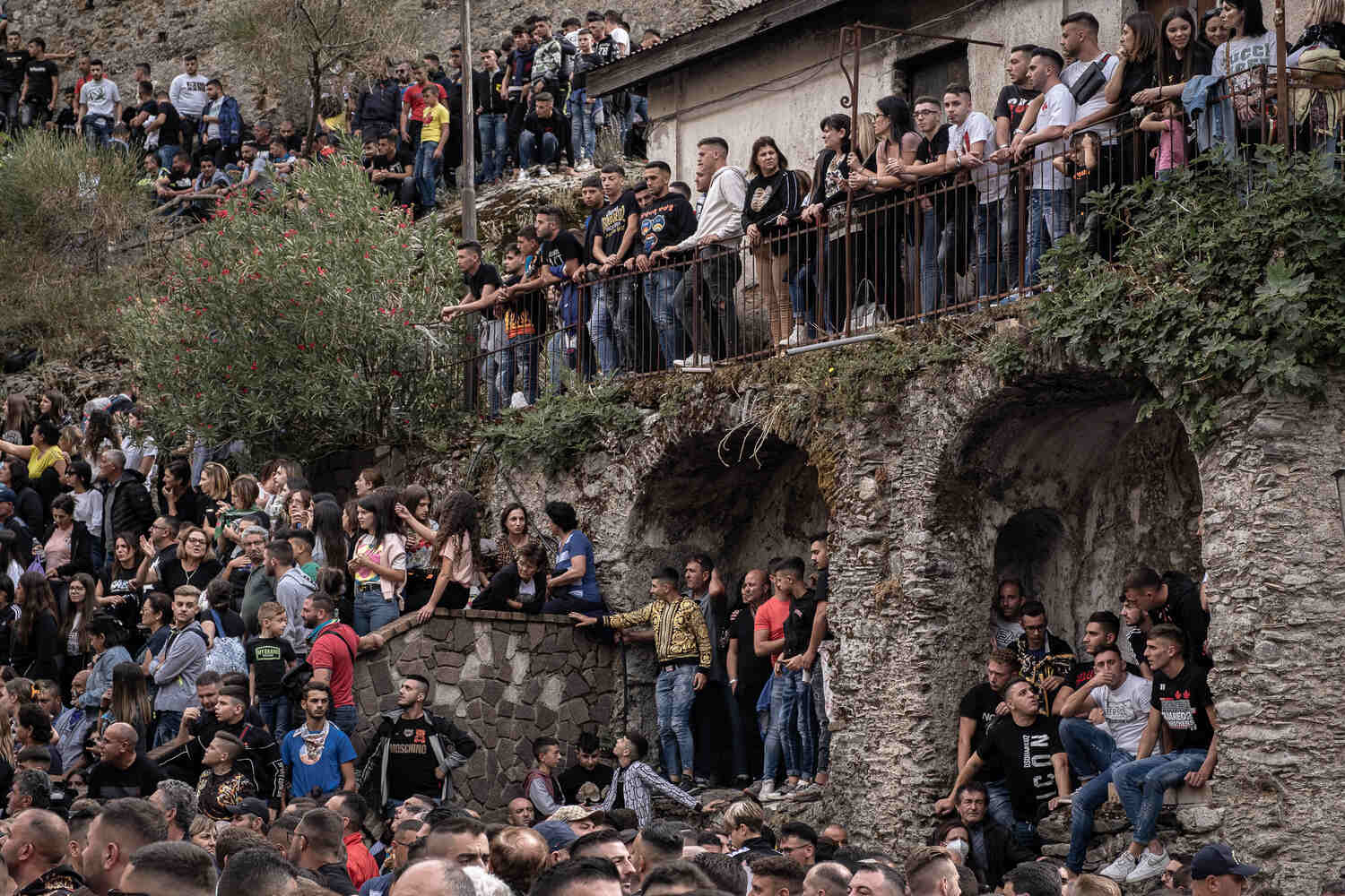 Hundreds of people, mostly young men, crowd railings and stairways looking down at something out of the frame.In the center of the frame is a young man wearing a very vivid gold jacket.