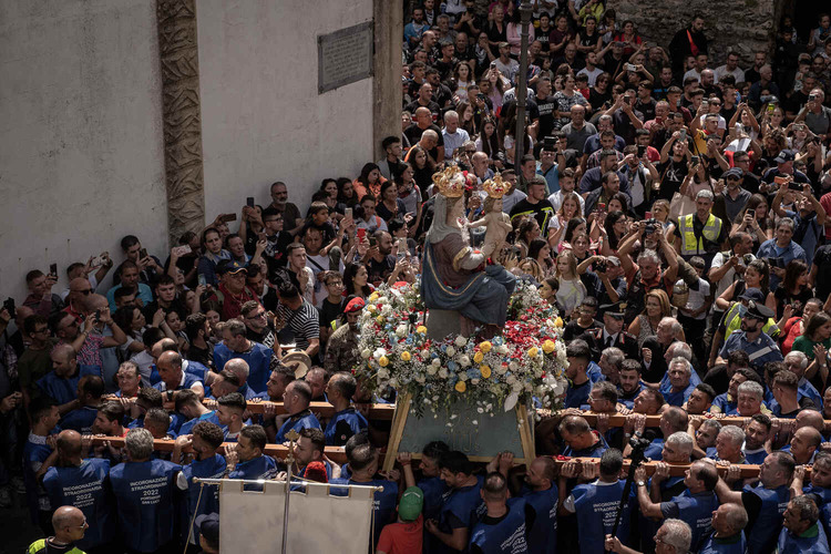 The statue of the Madonna and Child, with each wearing a crown, is carried on a wooden frame by more than 30 people, among a crowd of onlookers who are watching and taking photos with their phones.