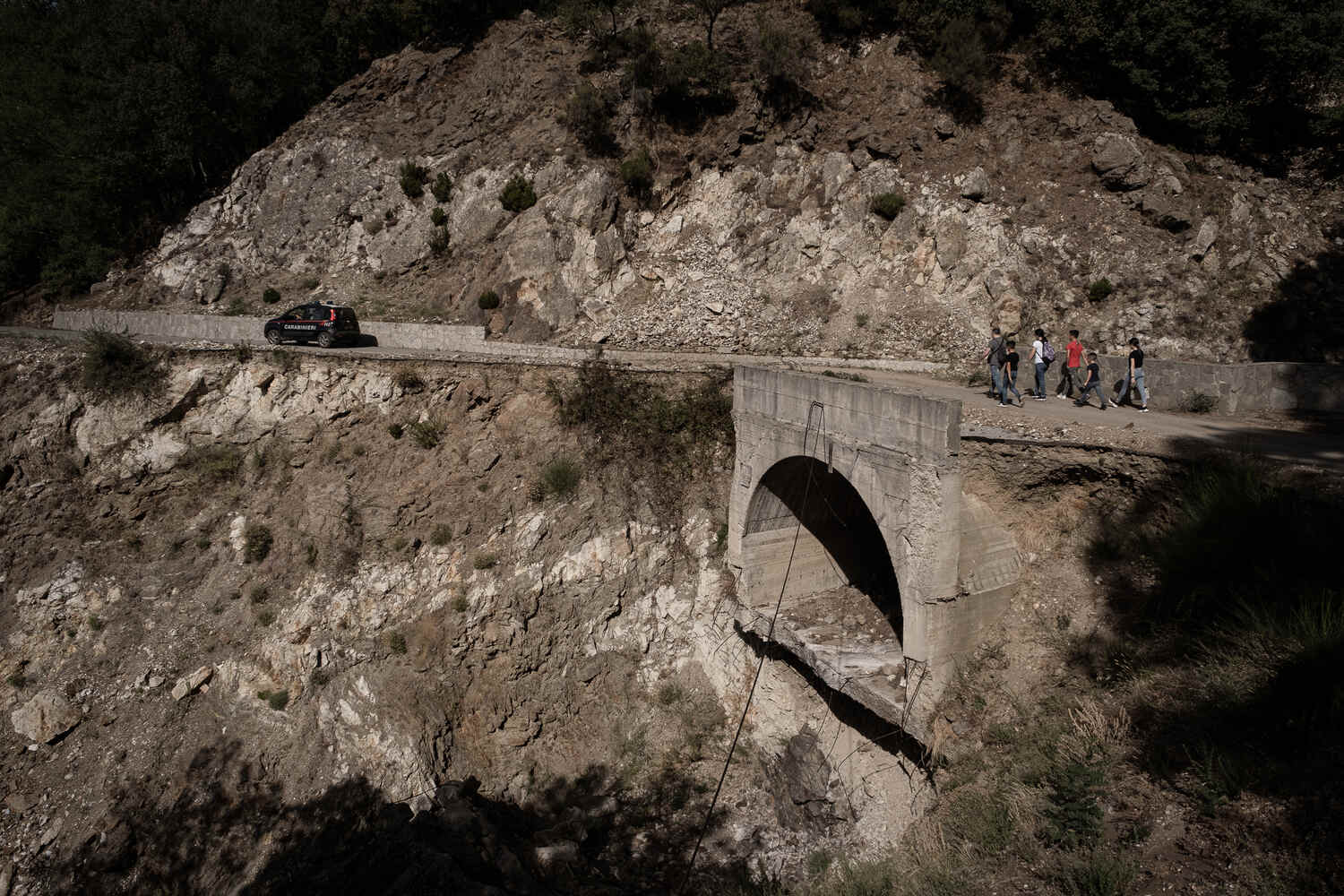 A mountain road with an Carabinieri police car seen driving off and a few pedestrians walking some way behind it.