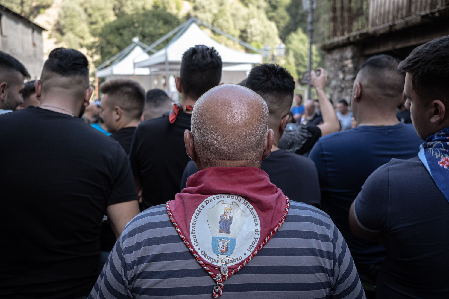 A procession moving forward outside seen from the back. The character in the center of the frame is wearing a red scarf which reads 'Confraternità Devoti della Madonna di Polsi – Campo Calabro'