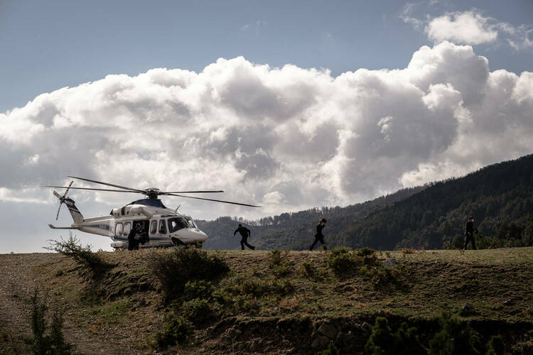 A helicopter perched on a green ridge under a blue sky, with 2 men walking towards it.