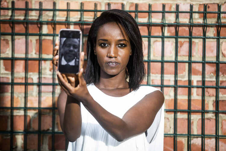 Carine Kanimba in front of a brick wall looking at the camera, holding a cellphone with the screen turned towards the camera. On the screen is a black and white photo of a man.
