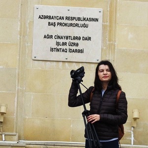 Sevinj Vaqifqizi holding a video camera in front of a building with a sign reading Prosecutor General's Office of Azerbaijan